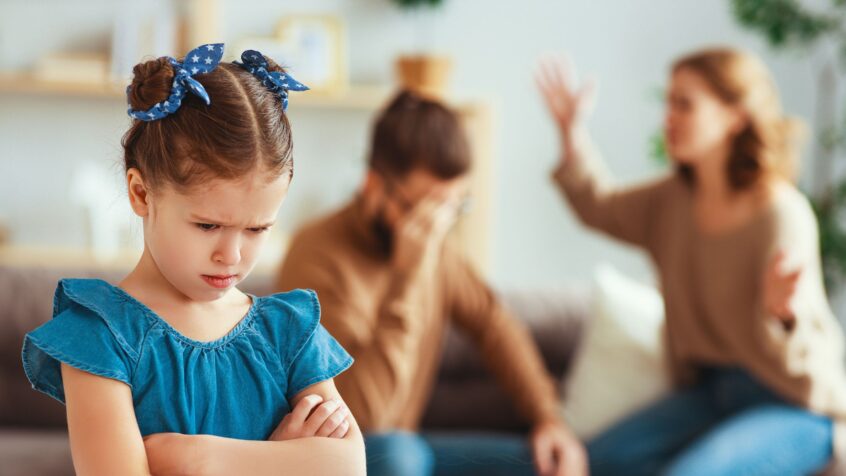 Petite fille contrariée au premier plan, avec ses parents discutant en arrière-plan, flous